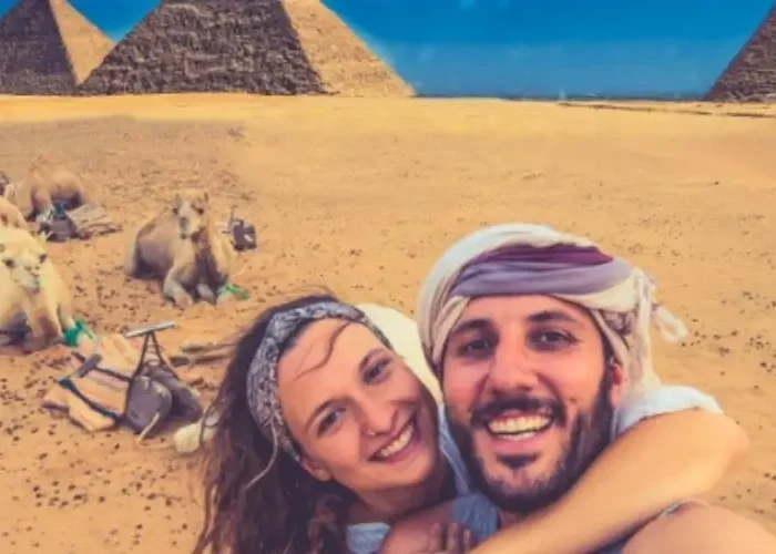 A smiling couple taking a selfie in front of camels and the Giza pyramids while traveling in Egypt in December.