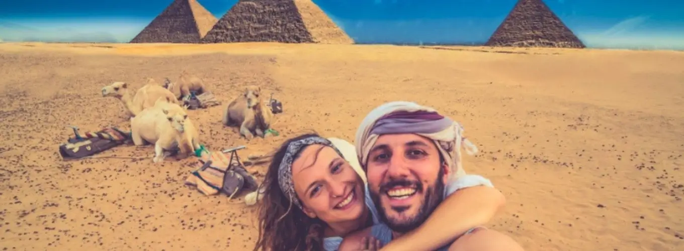 A smiling couple taking a selfie in front of camels and the Giza pyramids while traveling in Egypt in December.