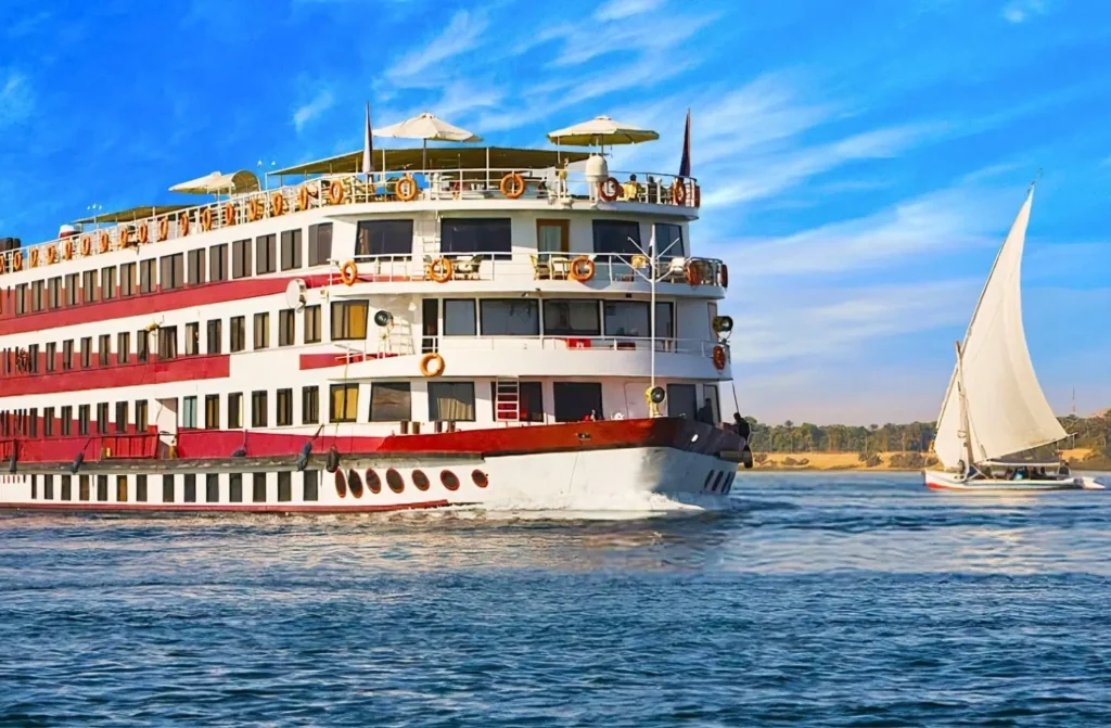 Large red-and-white Nile cruise ship sailing alongside a traditional sailboat, illustrating the visual contrast in a Nile Cruise vs Dahabiya comparison.