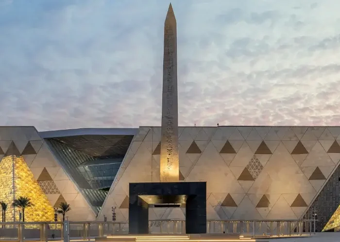 The illuminated exterior facade and Hanging Obelisk at the main entrance plaza of the Grand Egyptian Museum.