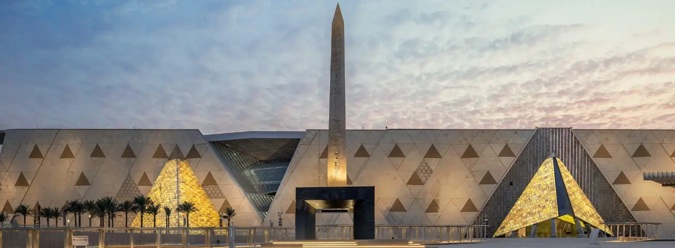 The illuminated exterior facade and Hanging Obelisk at the main entrance plaza of the Grand Egyptian Museum.